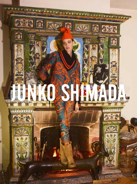 An adult woman models a patterned suit and boots in front of an ornate fireplace with burning logs in an indoor environment.