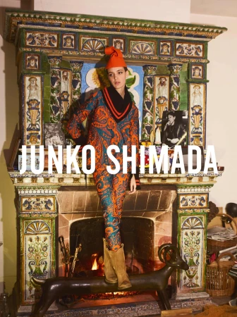 An adult woman models a patterned suit and boots in front of an ornate fireplace with burning logs in an indoor environment.