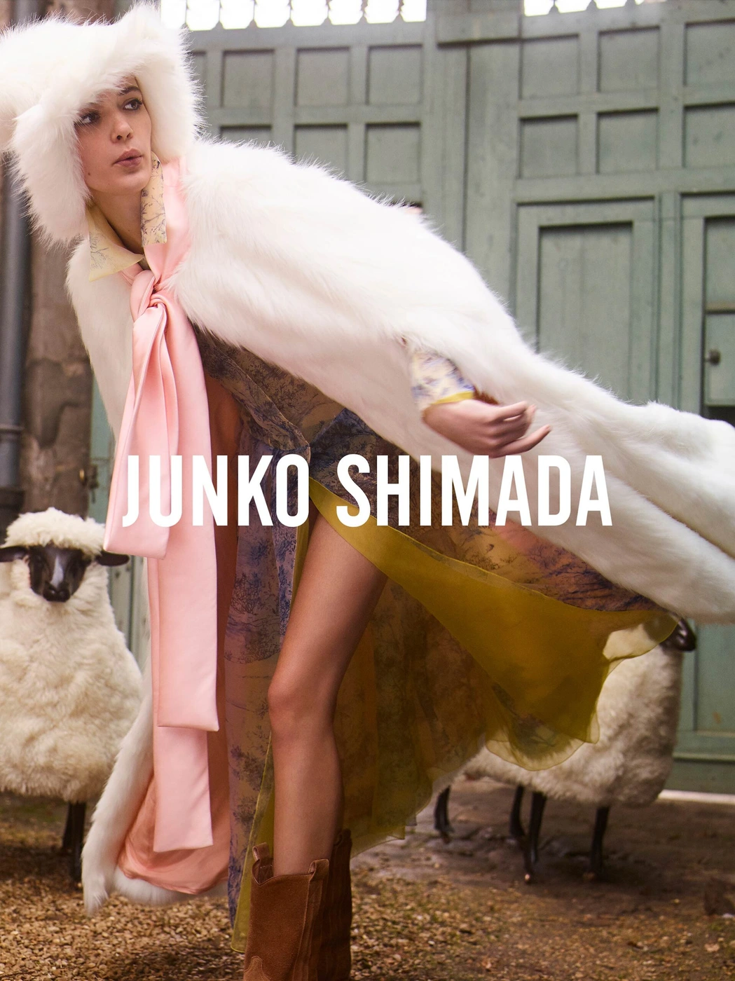 Model in white faux fur hooded cape and patterned dress standing near sheep outdoors by a wooden door