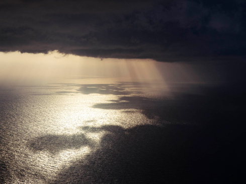 Dramatic aerial view of sunbeams over a dark ocean under a stormy, cloudy sky, creating a contrasting and moody seascape.