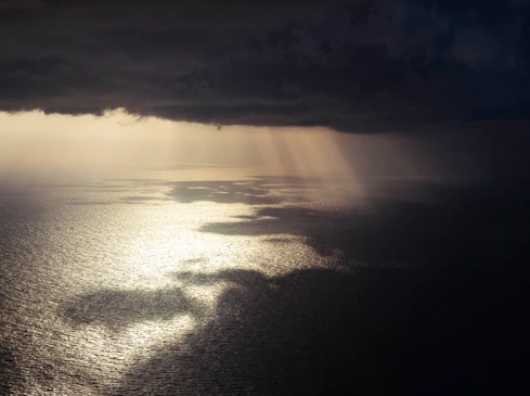 Dramatic aerial view of sunbeams over a dark ocean under a stormy, cloudy sky, creating a contrasting and moody seascape.