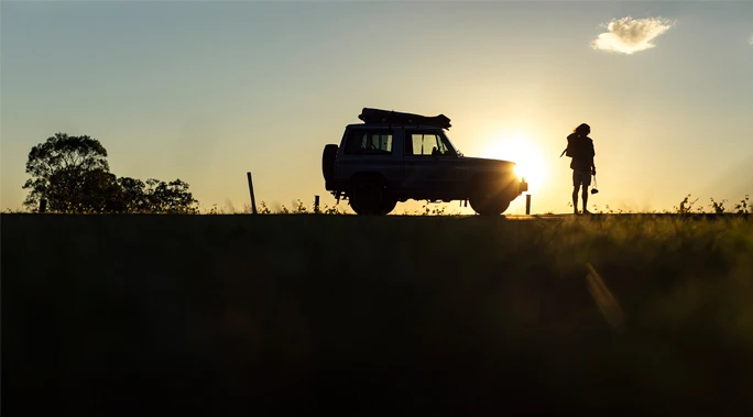Silhouette of an adult near a car at sunset with a large bright sun star, trees, and grassy fields in the background.