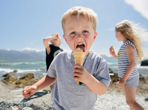 A young boy with blonde hair eats ice cream on a rocky beach with other children and mountains in the background on a sunny day.