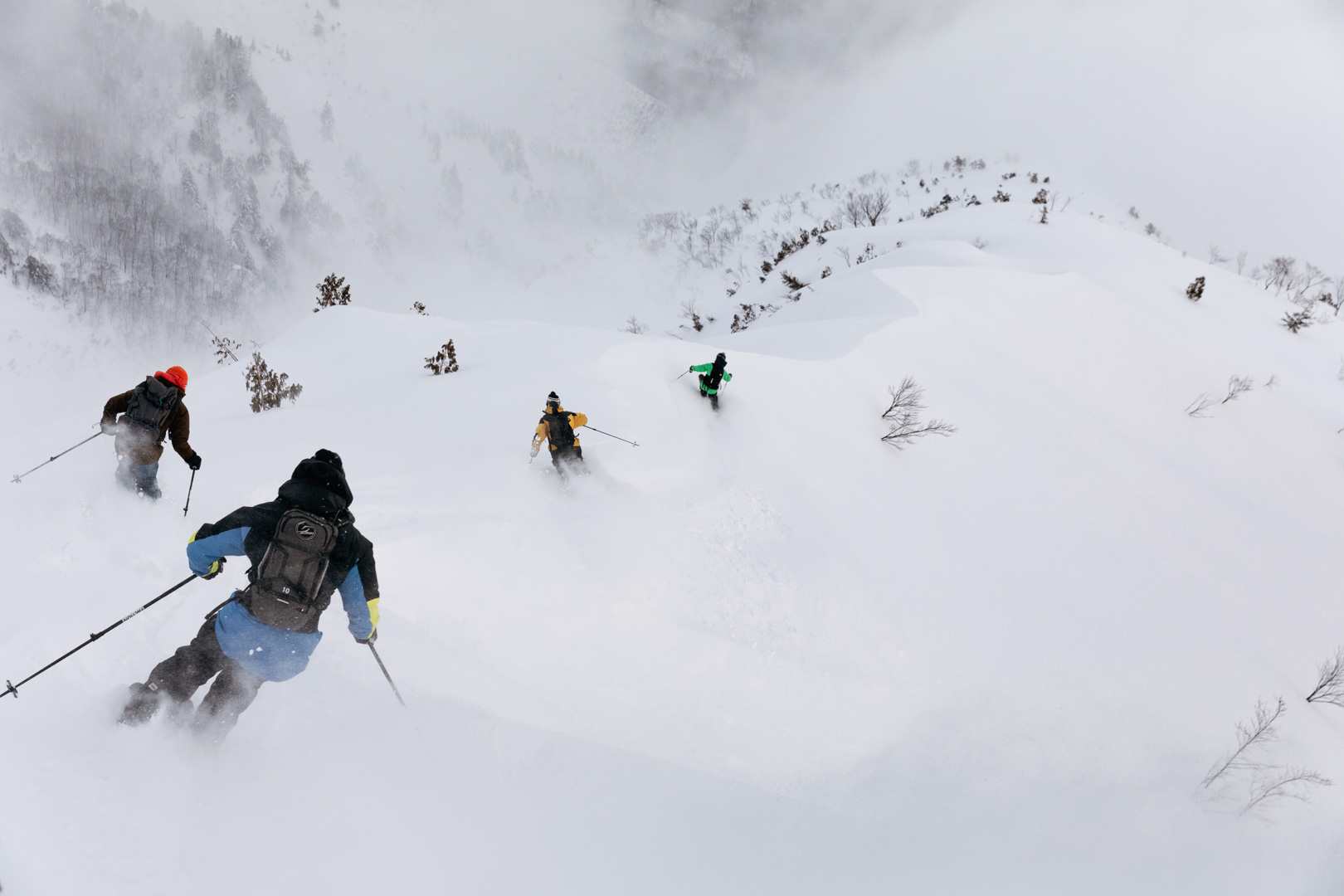 Four skiers descend a snowy mountain slope on a cloudy day, using ski poles to navigate the terrain covered with fresh powder snow.