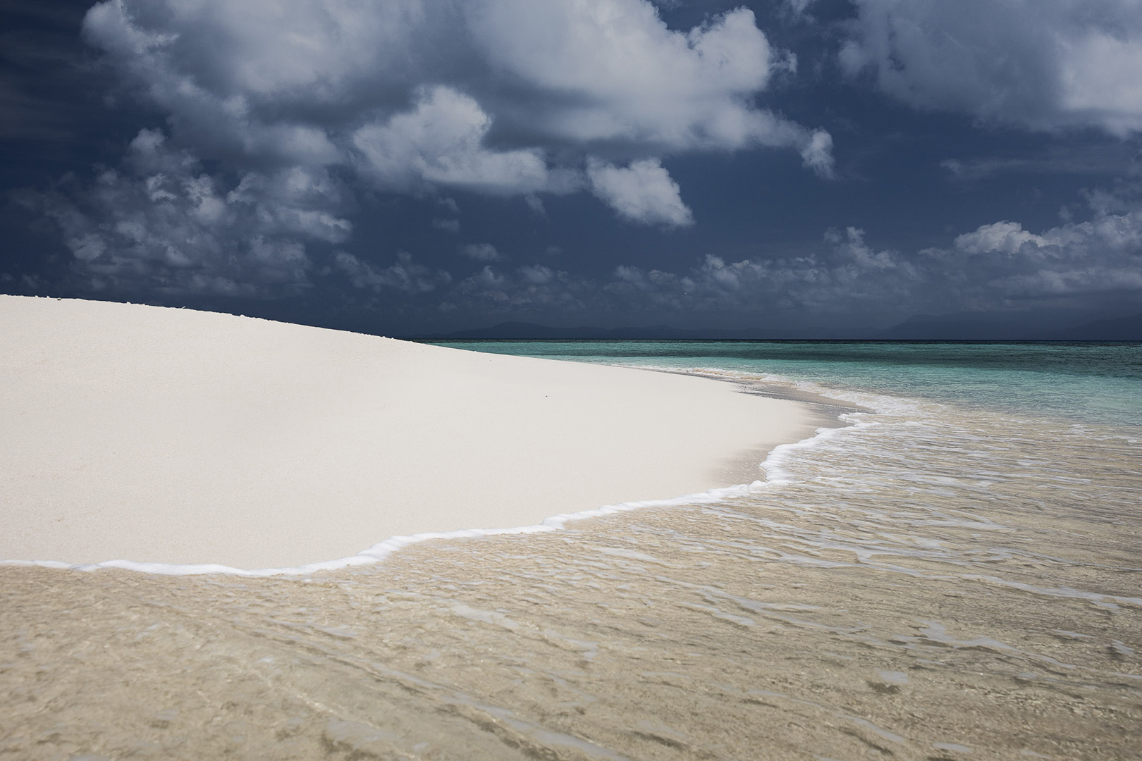 Island seascape featuring a white sandy beach meeting the turquoise ocean under a dramatic sky filled with dark and white clouds.