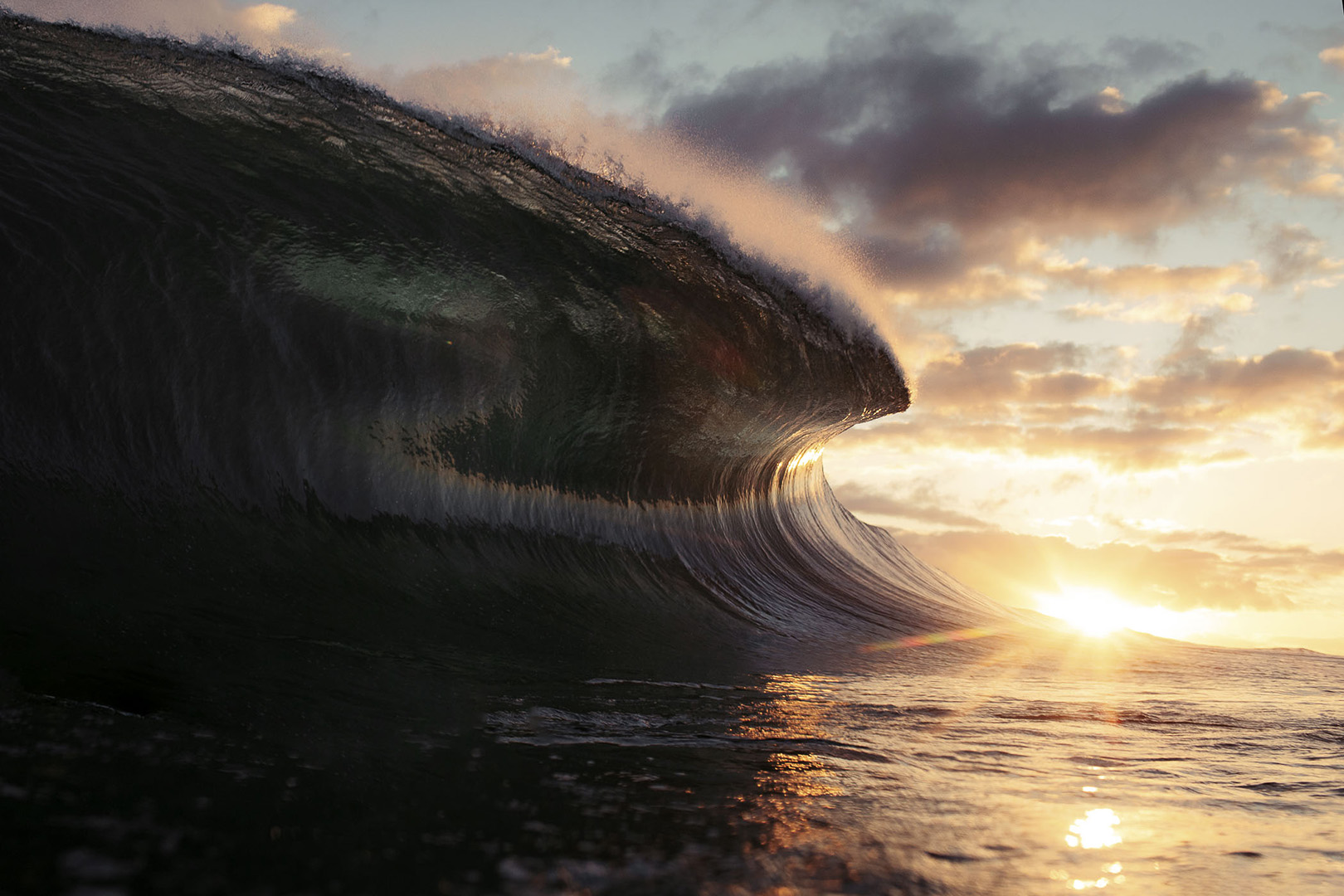 A large wave crests in the ocean at sunset, with light reflecting off the water and sky, showcasing nature's power and beauty.