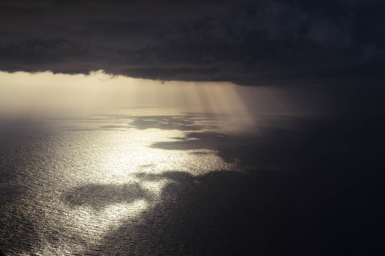 Moody seascape featuring dark clouds above the water's surface, with sunrays piercing through to illuminate the sea below.