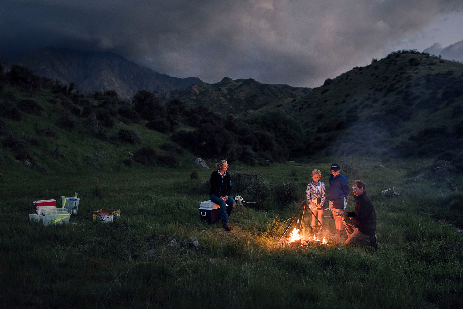 Family gathered around a campfire in a grassy field at dusk, surrounded by mountains. Woman sits on cooler, two children and man tend fire.