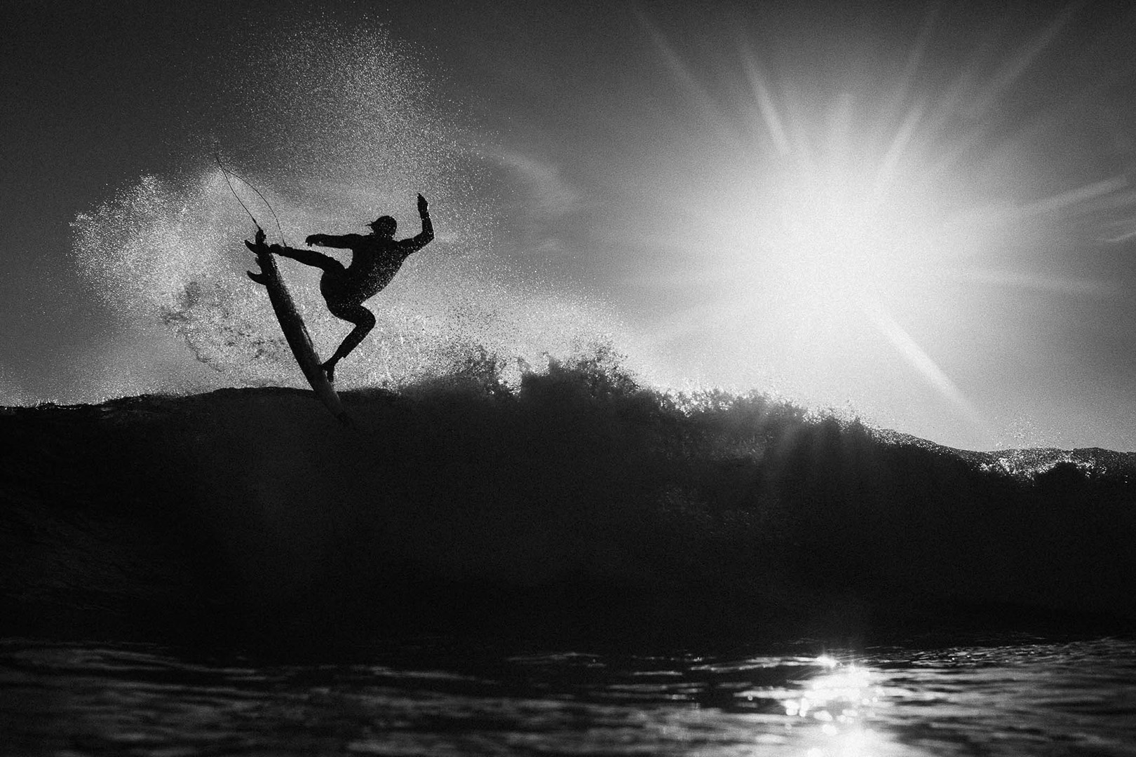 A silhouetted surfer catches air off a wave at sunset, captured in a black-and-white photograph showcasing the sport of surfing.