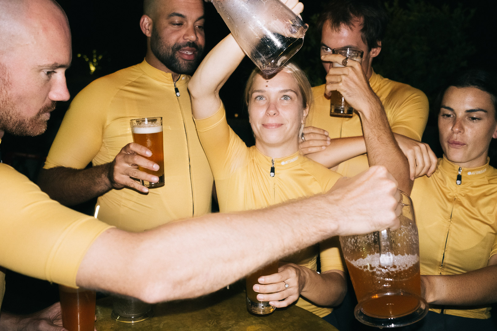 Group of six people in yellow shirts holding beer glasses and a pitcher in an indoor setting with dark background.