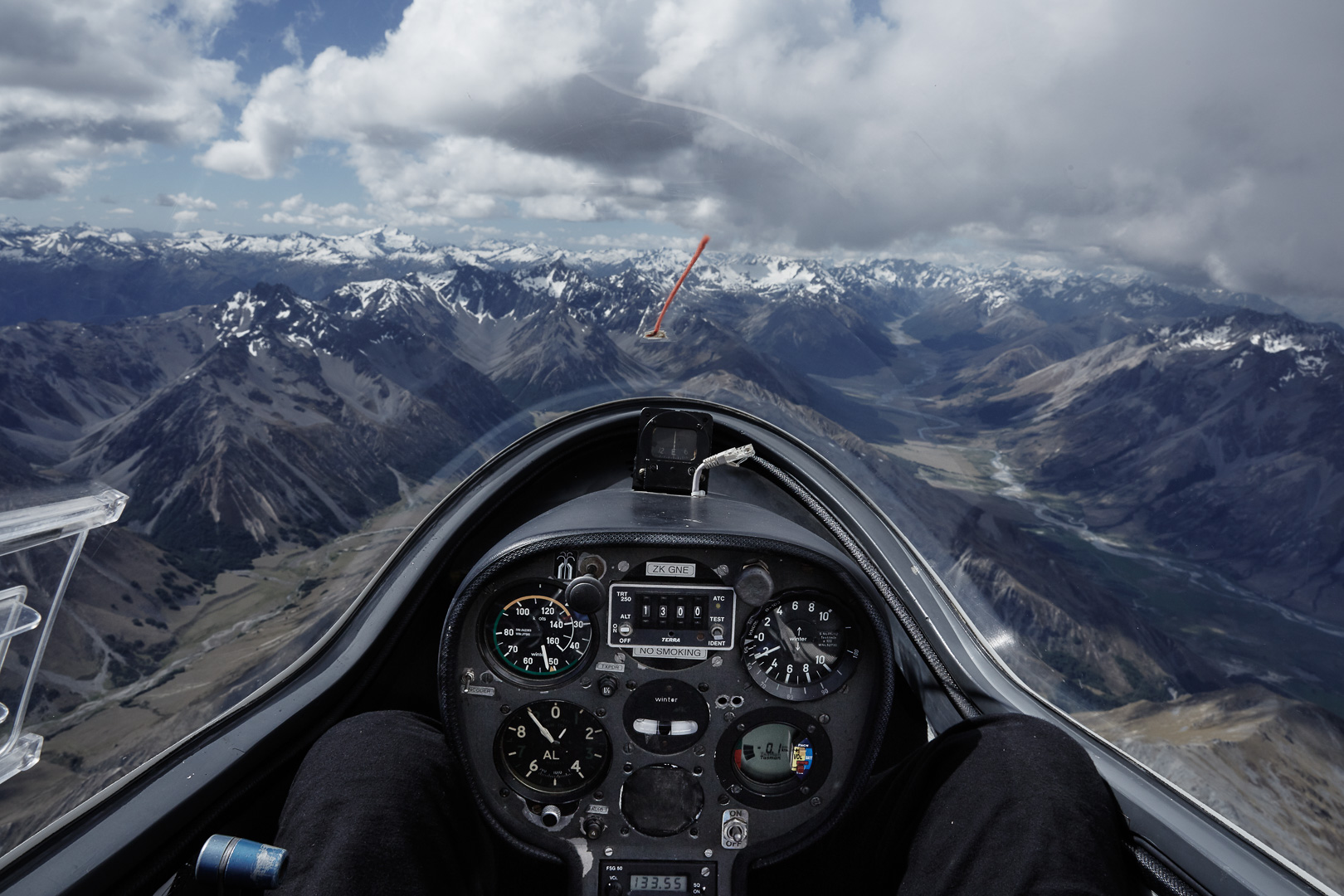 View from a glider cockpit over a mountain range, showcasing flight instruments and snow-capped peaks in the distance on a clear day.