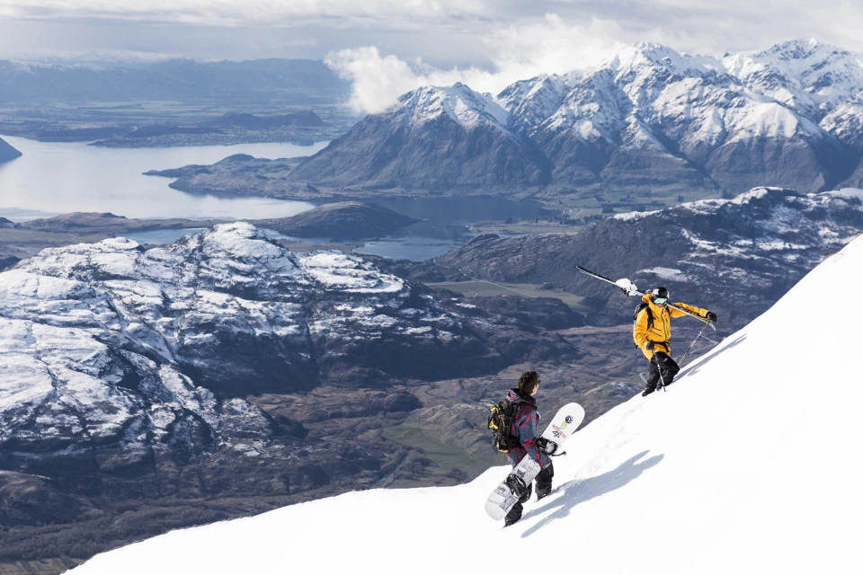 Two snowboarders and a skier hike up a snow-covered mountain slope on a sunny day with a lake and snow-capped mountains in the background.