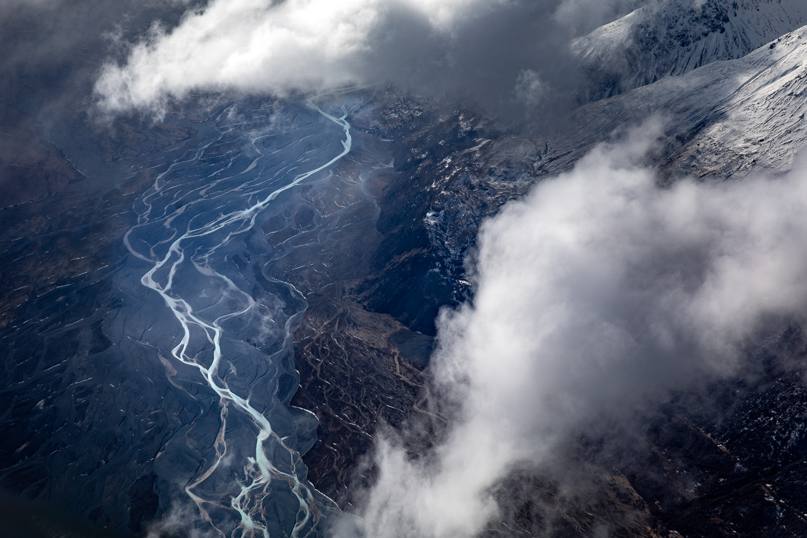 Aerial view of a braided river system winding through a mountain valley landscape with snow-capped peaks and low-lying cloud cover.