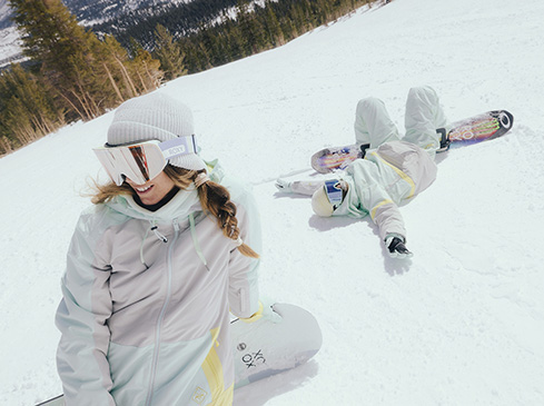 Two female snowboarders pose on a snowy slope with a backdrop of pine trees and a bright, overcast sky. One lies on her back.