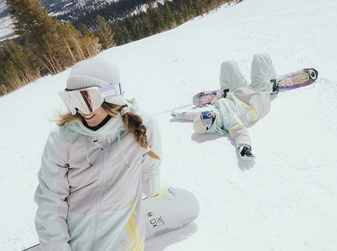Two female snowboarders pose on a snowy slope with a backdrop of pine trees and a bright, overcast sky. One lies on her back.