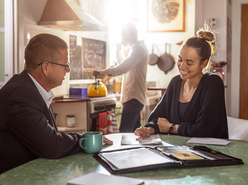 Two adults at a table with documents and coffee, while another person stands in the kitchen area in the background, bathed in bright sunlight.