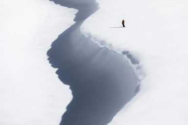 Aerial view of a lone skier standing near a massive snow crevasse on a vast white snowfield.