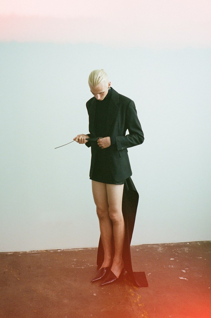 Fashion shot of a fair-skinned adult male with braided blond hair in a black blazer, turtleneck, and skirt standing in front of a white wall.
