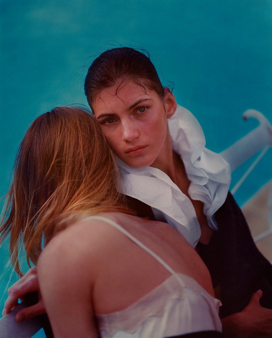 Two women, one in white, pose intimately against a blue background, with one looking directly at the camera in a fashion-style shot.