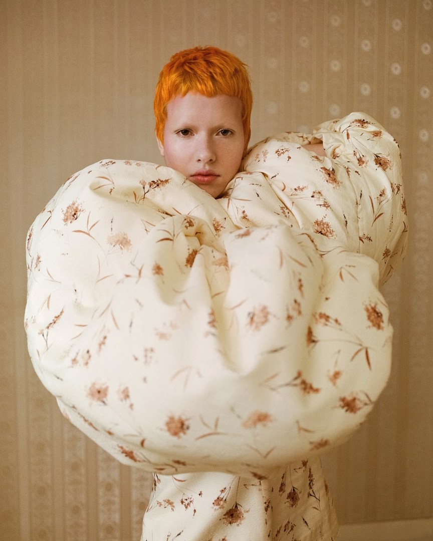 Portrait of a person with bright orange hair wearing a floral pattern dress, posing in an indoor setting with neutral background.