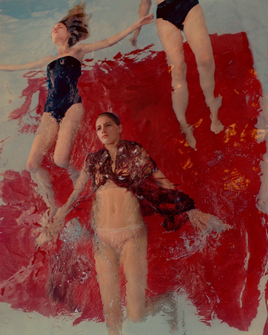 Three women in swimsuits and casual wear are submerged in rippling water with a red and white pigment effect on the water's surface.