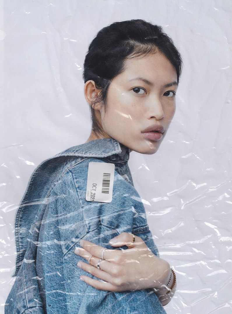 Portrait of a fashionable young woman wearing a denim jacket with a tag, posing against a wrinkled plastic backdrop in a studio setting.