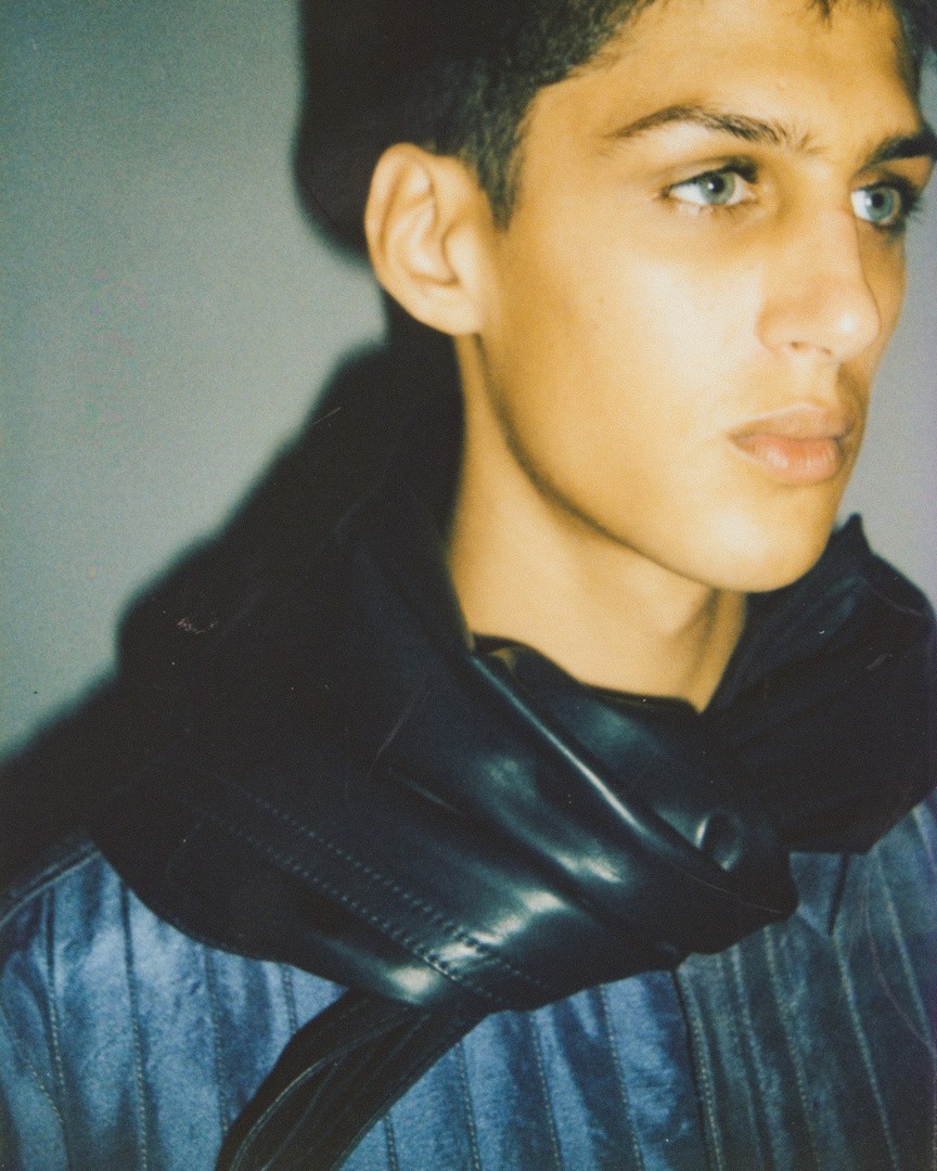 Portrait of a young adult with light eyes wearing a black scarf and blue and white striped shirt against a grey backdrop.