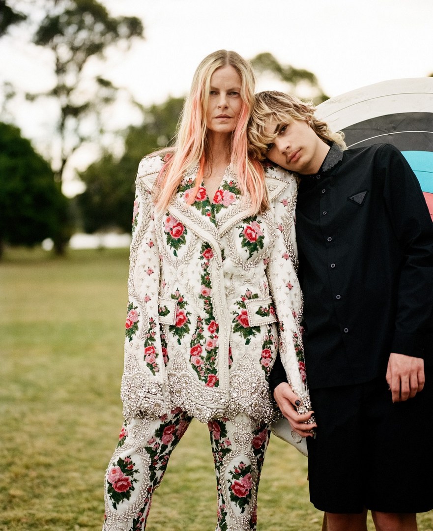 Two people pose in an outdoor setting. An adult woman with blond hair wears a floral suit while a younger person rests their head on her shoulder.