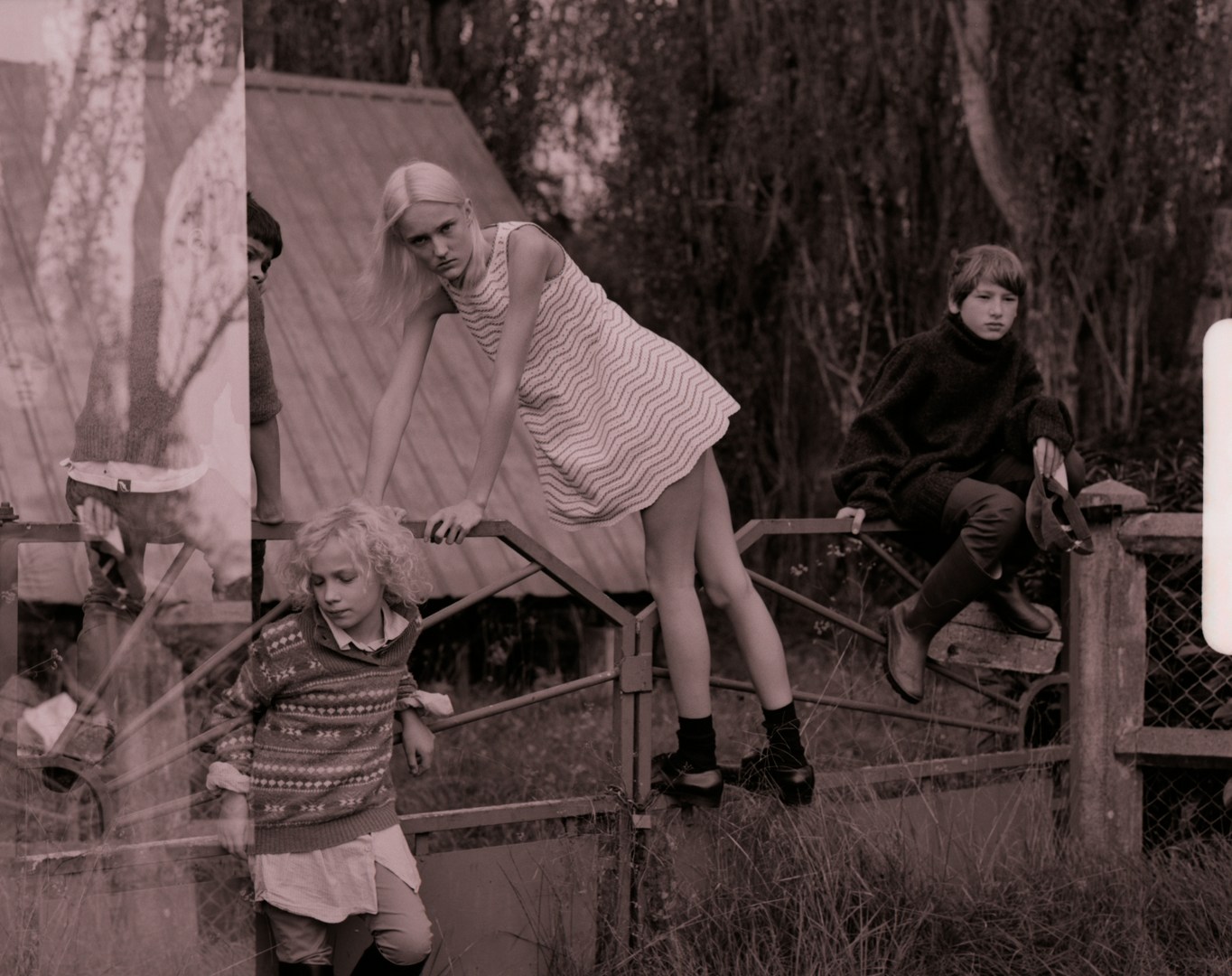 Three young people on a fence: a blonde girl in patterned dress stands on the fence, another sits on the fence, and another looks on.