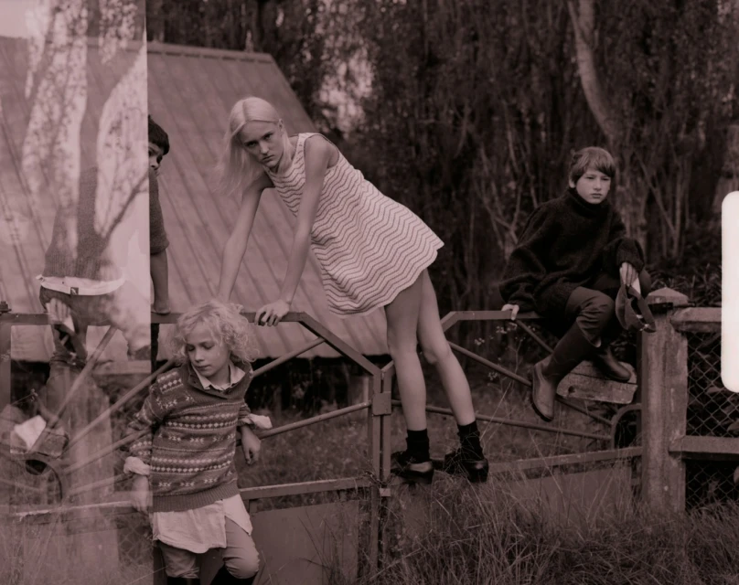 Three young people on a fence: a blonde girl in patterned dress stands on the fence, another sits on the fence, and another looks on.