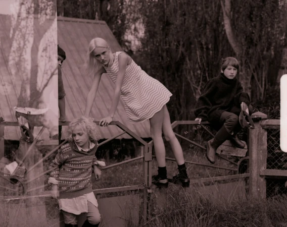 Three young people on a fence: a blonde girl in patterned dress stands on the fence, another sits on the fence, and another looks on.