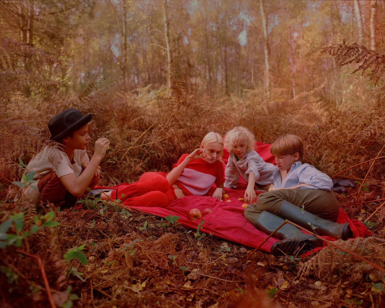 Four children gather on a red blanket in a forest clearing surrounded by ferns, enjoying a picnic with apples and late-afternoon sunlight.