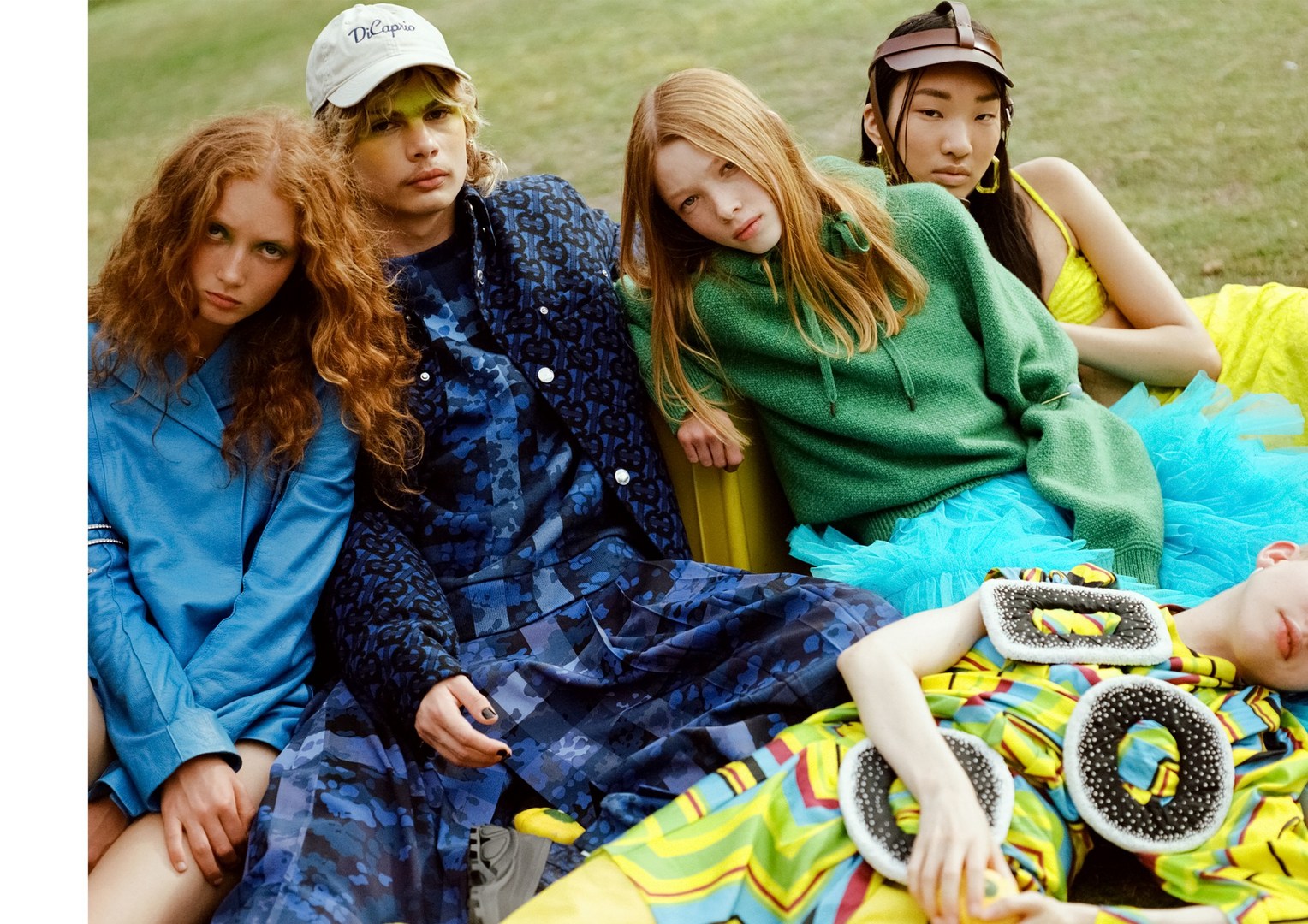 Group of five young fashion models in colourful streetwear sitting outdoors on a yellow bench in a grassy setting.