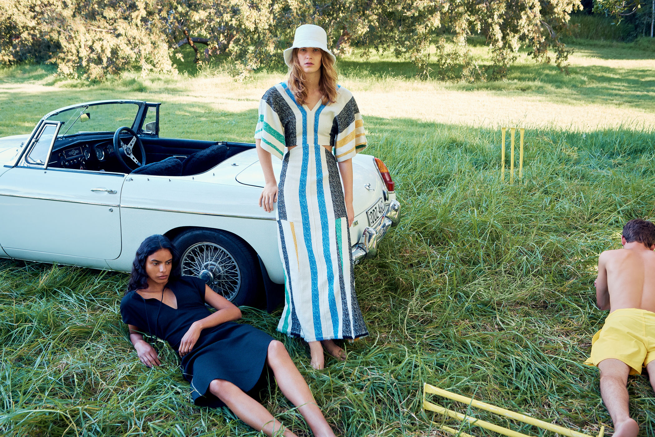 Two women and a man pose in a grassy field with a white convertible car and yellow cricket wickets under bright sunlight in a fashion editorial.