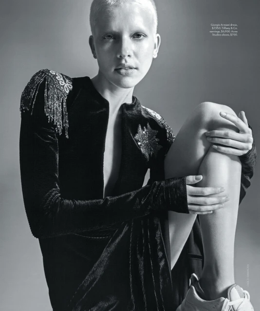 Monochrome image of a fashion model with an edgy haircut wearing a beaded black jacket, posing in a neutral grey studio environment.