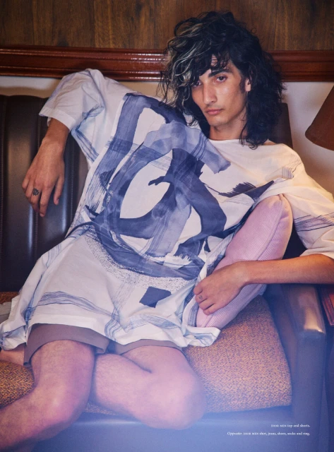 A young adult male model poses indoors, sitting in a chair, wearing a patterned white top and brown shorts in a fashion editorial style.