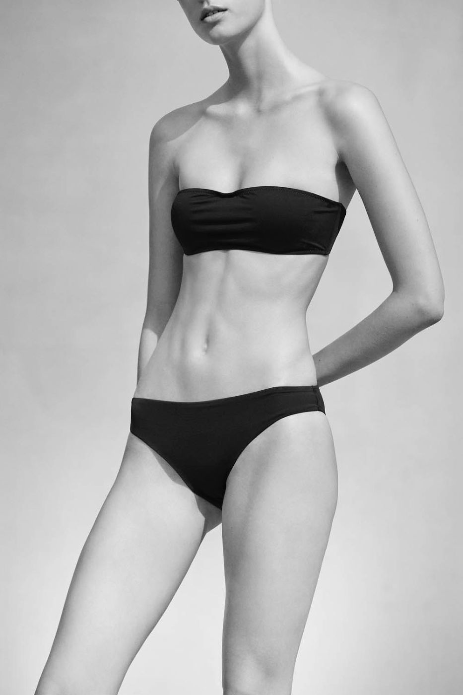Monochrome portrait of a young adult woman wearing a black two-piece swimsuit against a plain studio backdrop, showcasing simple swimwear fashion.