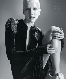 Model with shaved blonde hair wearing a black embellished velvet dress and sneakers against a grey backdrop.
