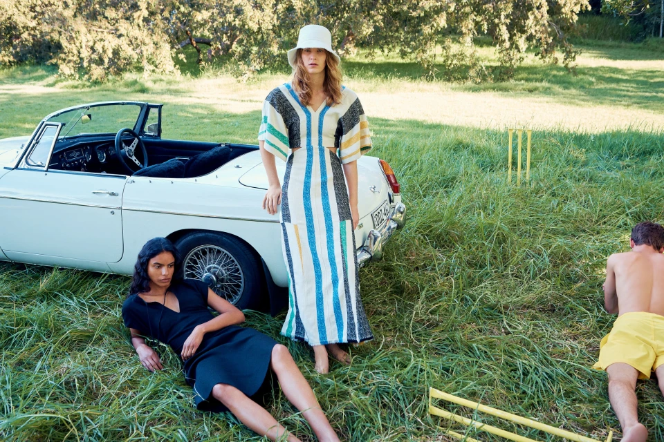 Two women pose on grass beside a white convertible car and cricket wickets under the sun. One man lies on the grass.