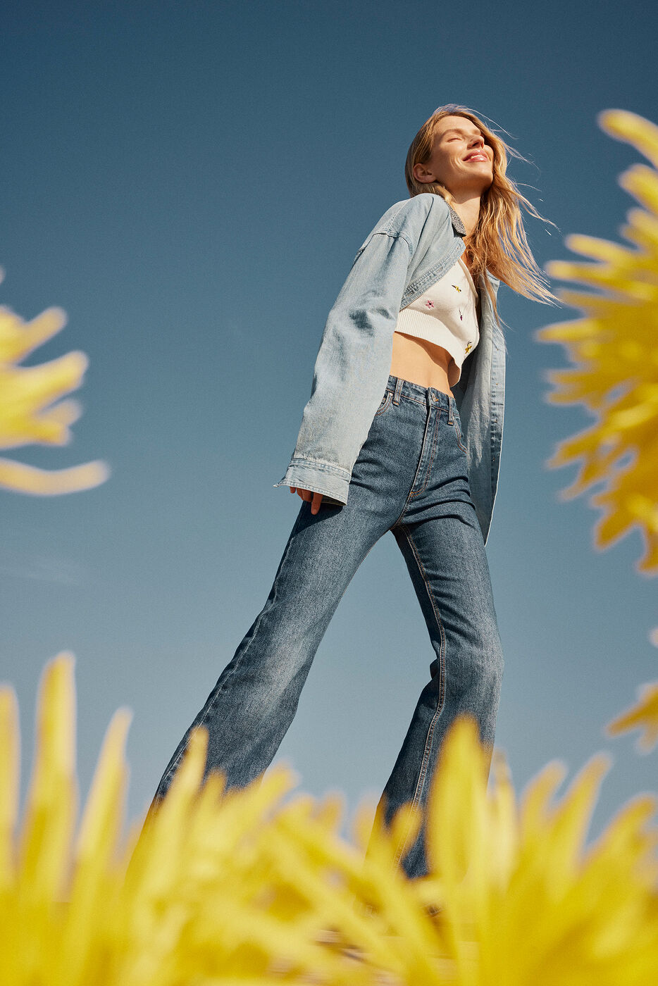 Low-angle shot of a smiling blonde woman wearing a denim shirt and flare jeans against a clear blue sky, framed by out-of-focus yellow flowers.