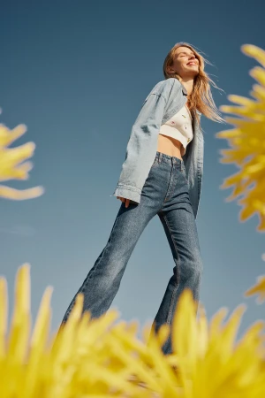Low-angle shot of a smiling blonde woman wearing a denim shirt and flare jeans against a clear blue sky, framed by out-of-focus yellow flowers.