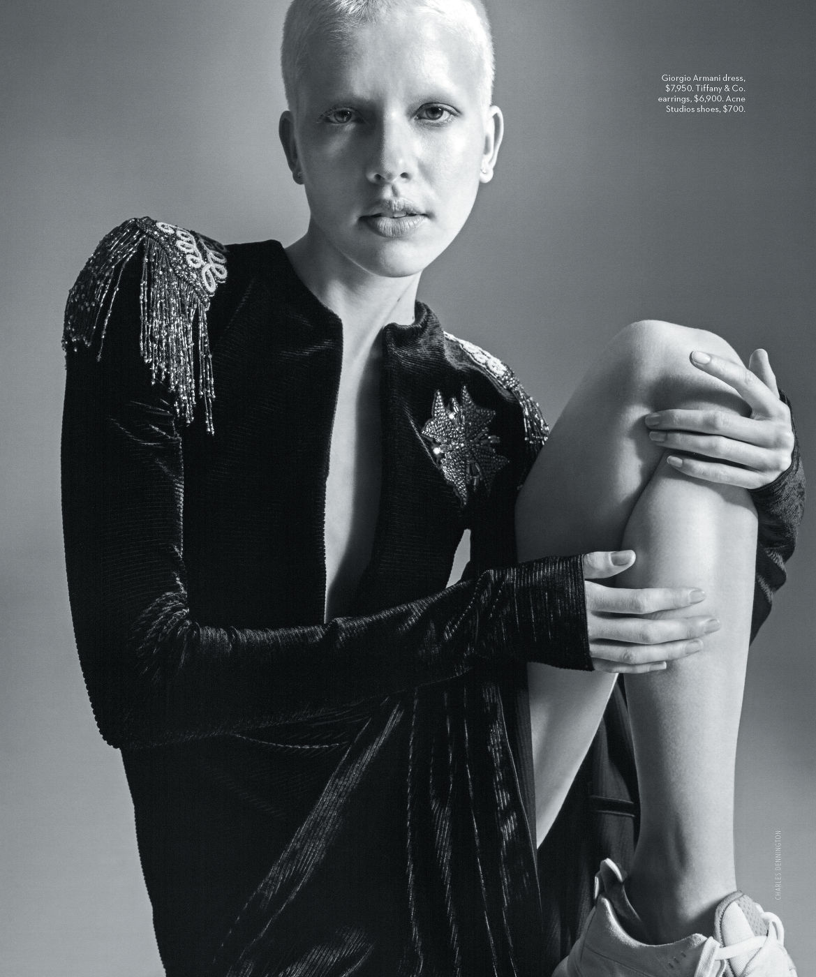 Monochrome fashion editorial portrait of a person with a shaved head wearing a dark embellished dress.