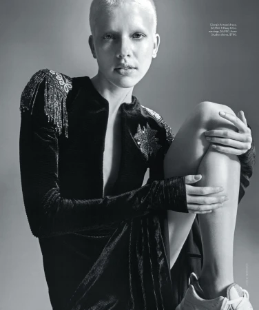 Monochrome fashion editorial portrait of a person with a shaved head wearing a dark embellished dress.