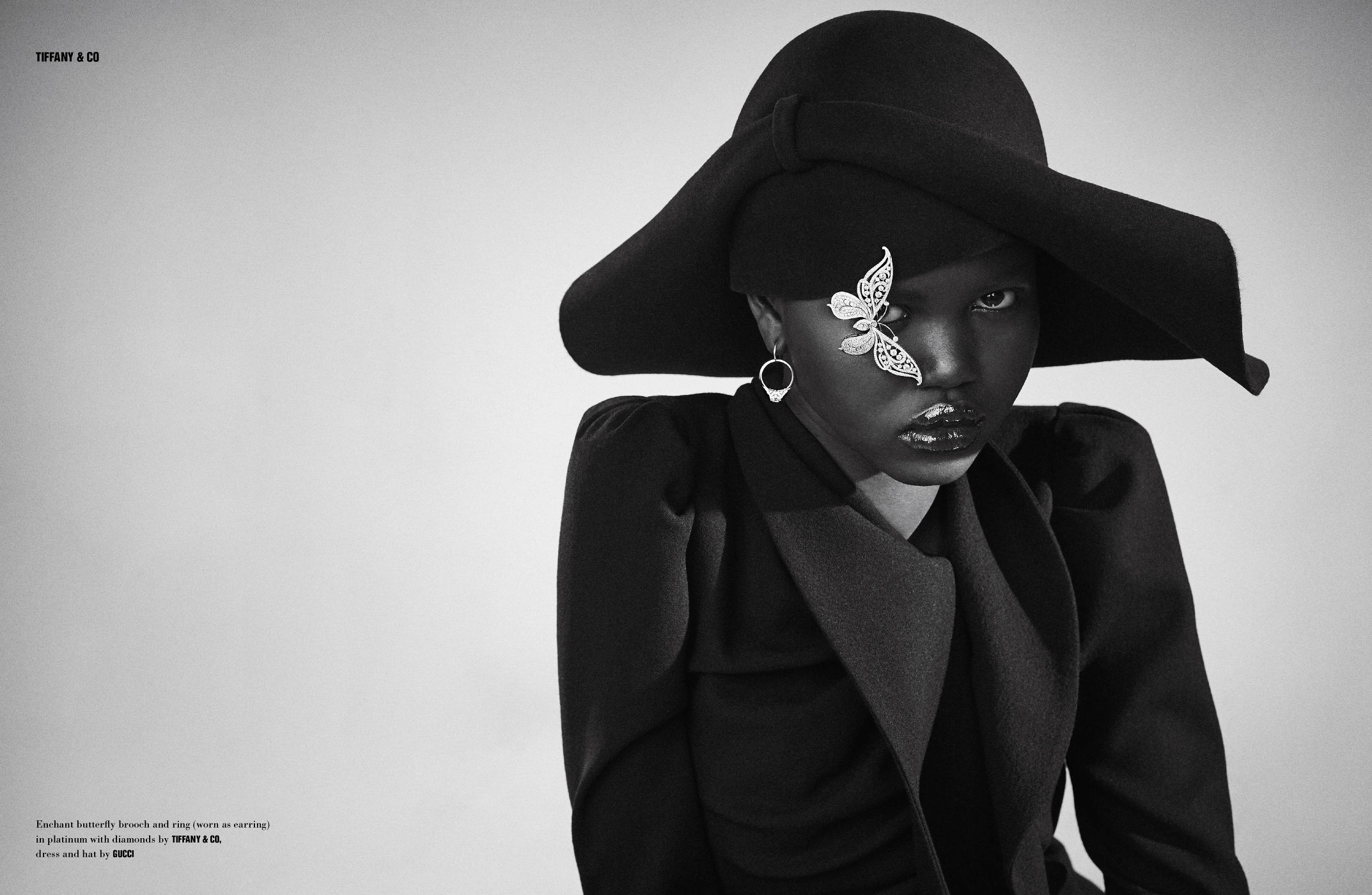 Monochrome fashion shot: Black woman wearing hat with butterfly brooch, statement earrings, and coat against a neutral backdrop.