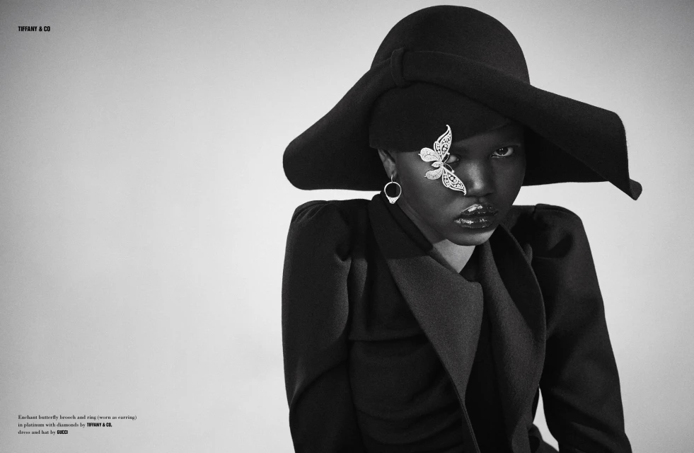 Monochrome fashion shot: Black woman wearing hat with butterfly brooch, statement earrings, and coat against a neutral backdrop.