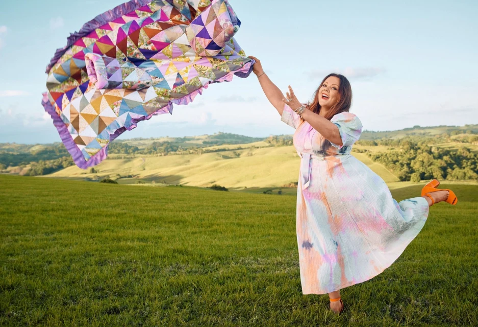 A smiling woman in a colorful dress stands in a grassy field holding a patchwork quilt, with rolling hills and blue sky in the background.
