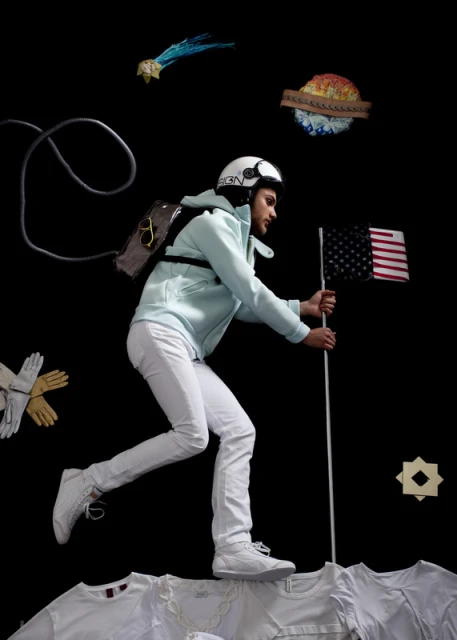 A man in white clothing strikes a pose with an American flag in a surreal, dreamlike scene against a stark black backdrop.