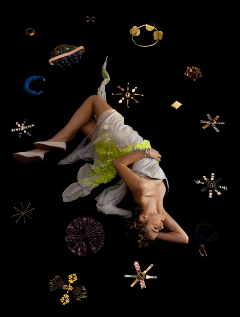 Artistic studio portrait of a woman lying amid floating fashion objects and jewelry against a black background in a unique editorial style.