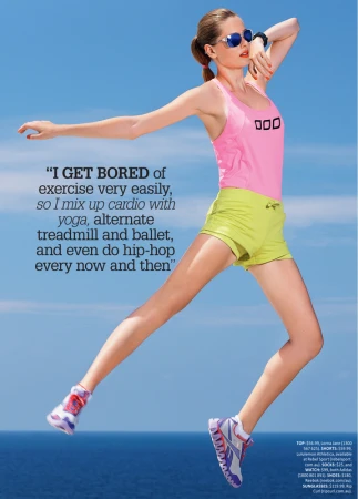 Woman in pink tank top and green shorts jumping mid-air against clear blue sky and ocean backdrop.