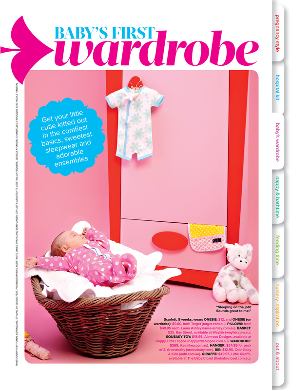 Baby girl sleeps in a wicker basket with baby clothing hanging on a pink door in a magazine editorial spread.